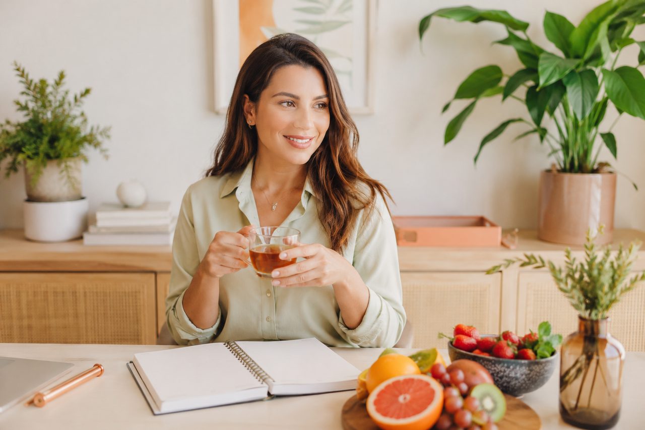 Mulher sorridente tomando chá em ambiente acolhedor, cercada de frutas e plantas
